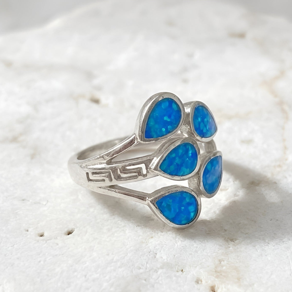 A silver ring with five teardrop-shaped blue opal inlays, displayed on a textured white stone surface.
