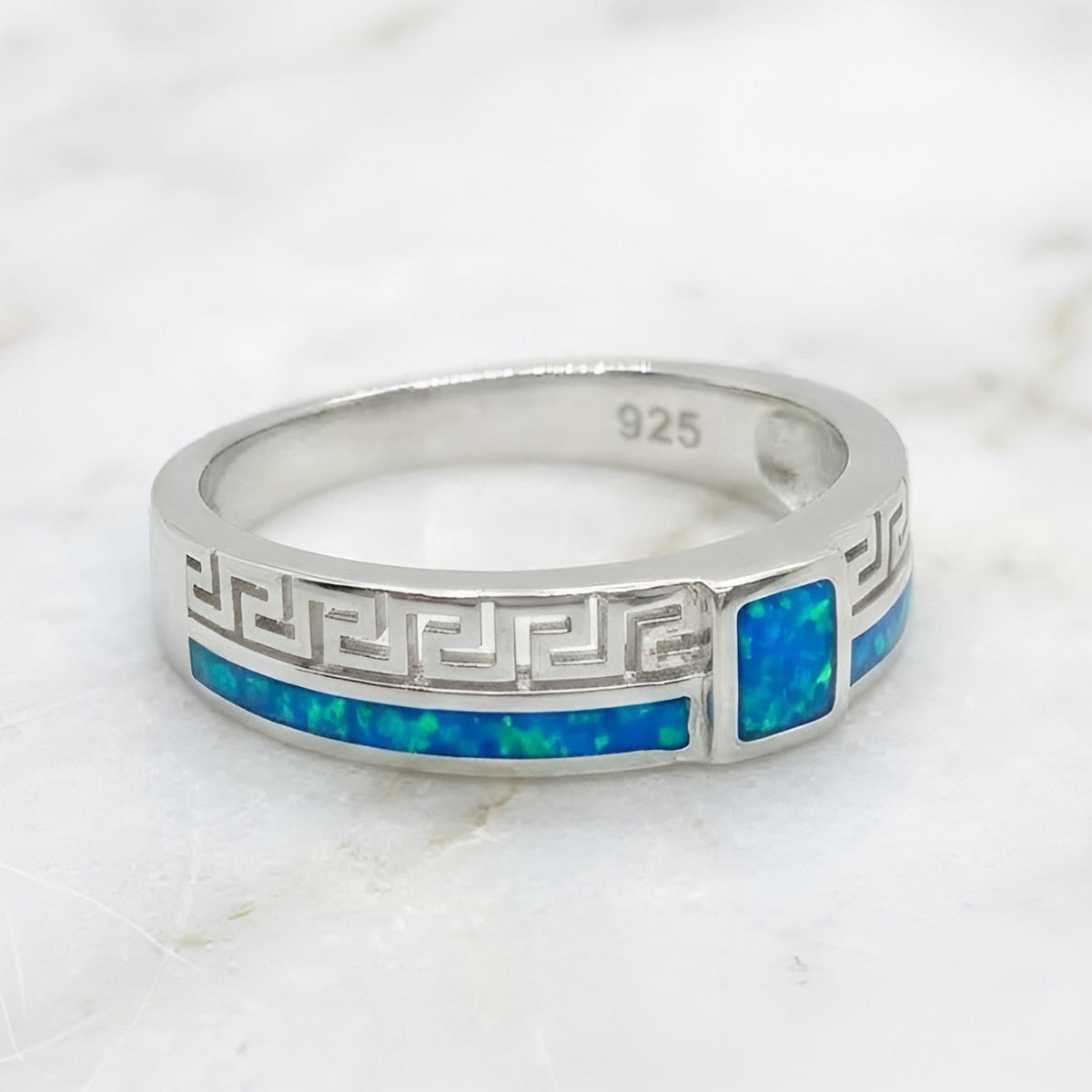A sterling silver ring with a Greek key pattern and a vibrant blue inlay, resting on a white marble surface.