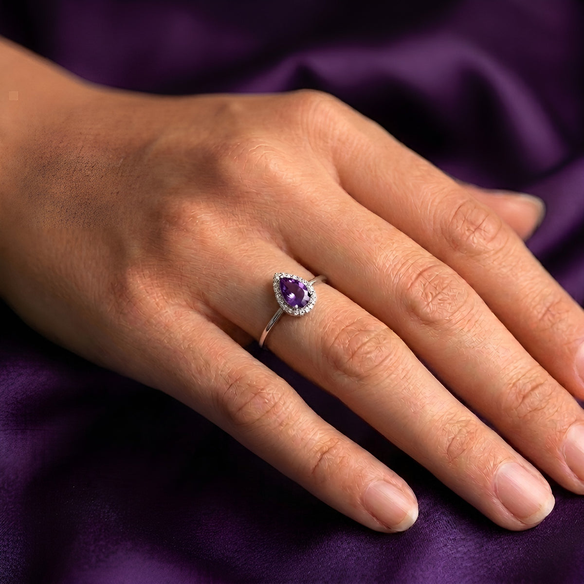 A close-up of a hand wearing a silver ring with a pear-cut purple crystal surrounded by small clear stones, set against a draped purple fabric background.