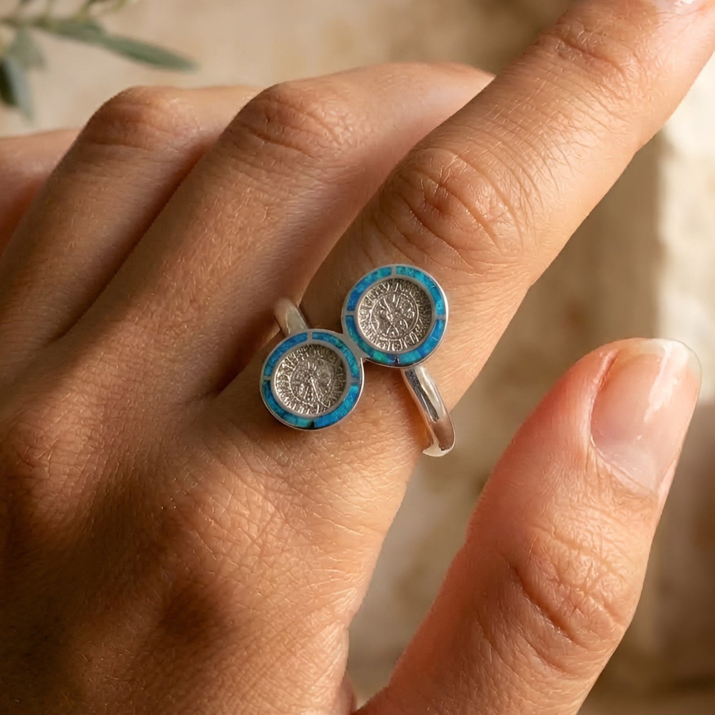 A close-up of a hand wearing a silver Phaistos Disc ring with two round blue and turquoise opal inlaid medallion-like tops, photographed in warm natural light against a softly blurred beige background.