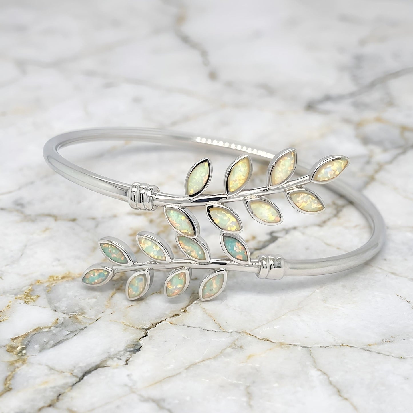 A silver bangle bracelet featuring two leafy branches inlaid with iridescent opal-like stones, displayed on a white marble surface.
