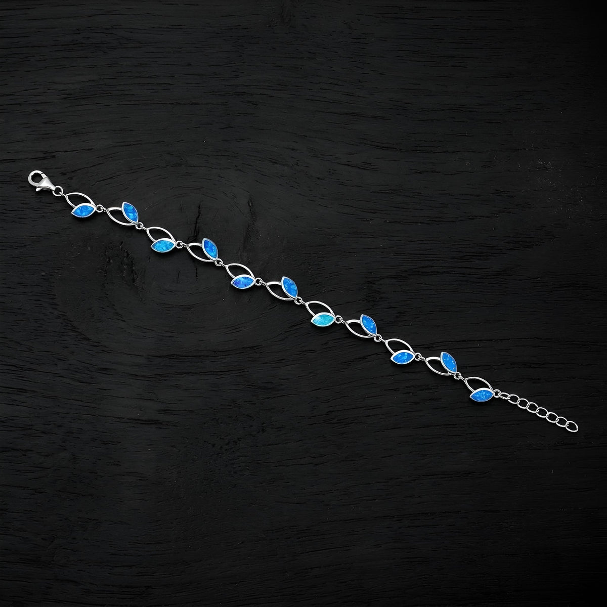 A silver link bracelet featuring a series of marquise-cut blue opal stones, displayed diagonally on a dark textured wooden surface.