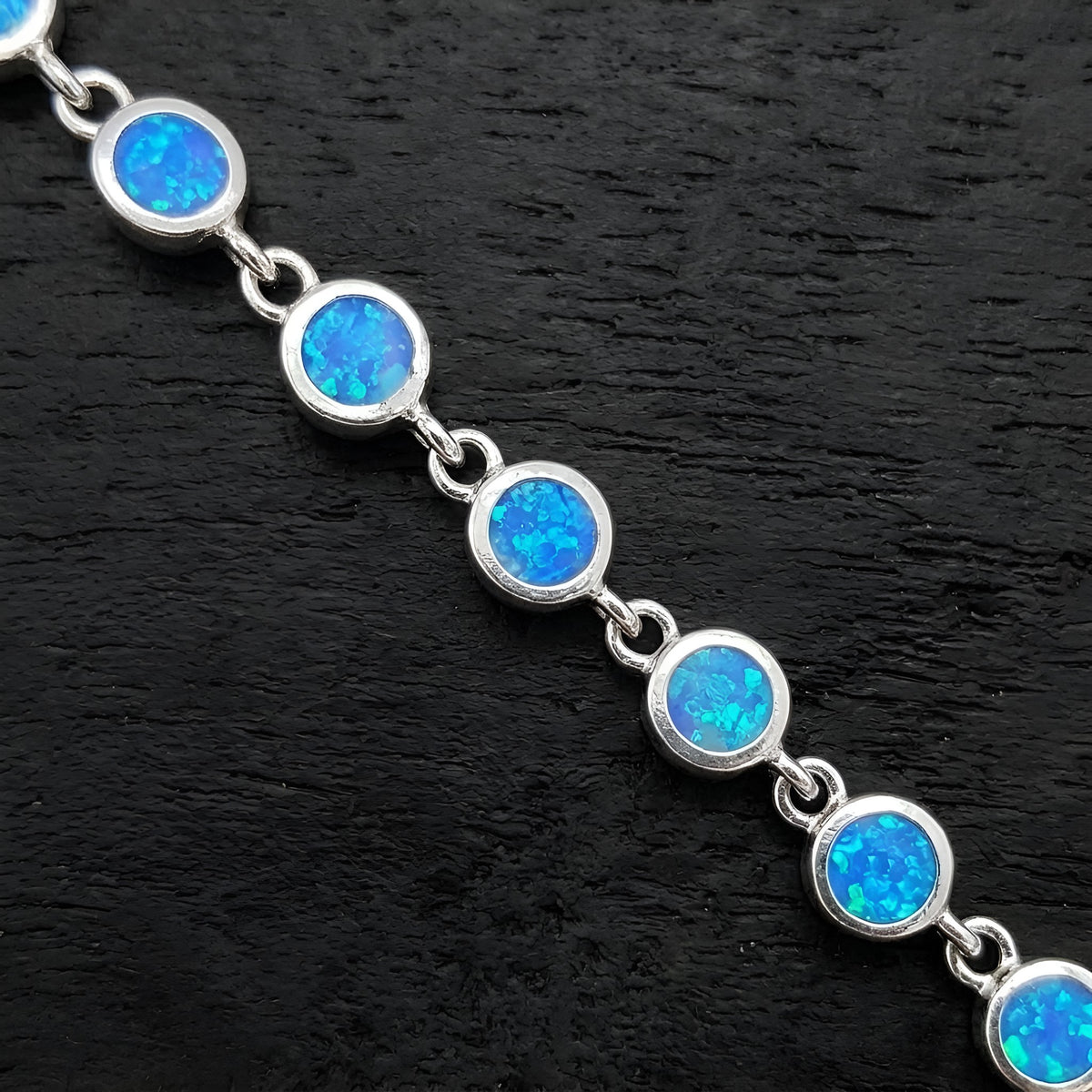 A silver chain bracelet featuring round blue opal gemstones, displayed diagonally on a dark textured background.