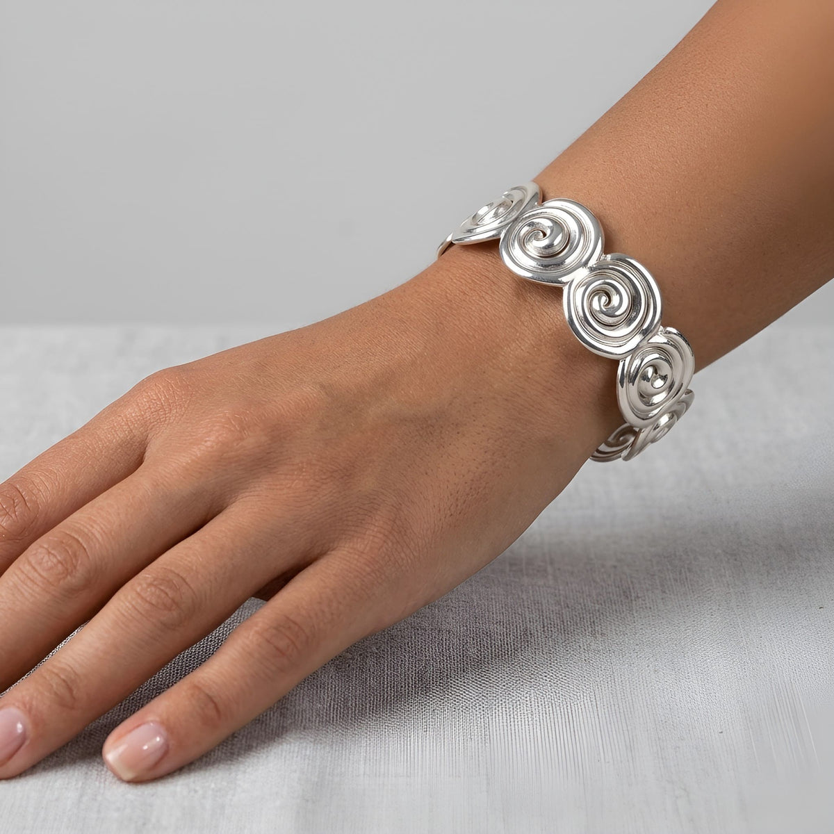 A close-up of a person's hand and wrist wearing a wide silver spiral cuff bracelet, resting on a light gray textured surface.