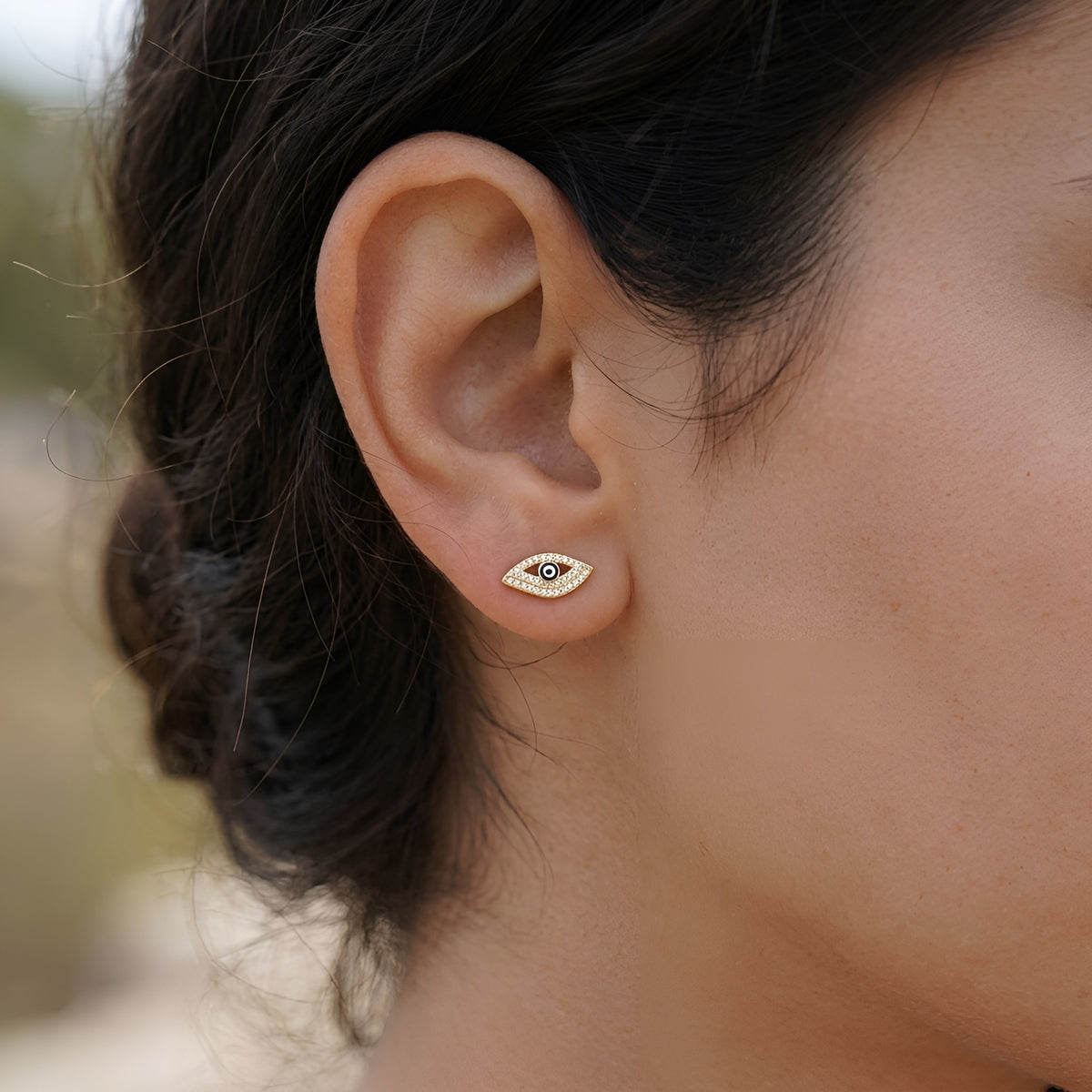 A close-up of a person's ear wearing a small gold eye-shaped stud earring with a  dark blue evil eye detail.