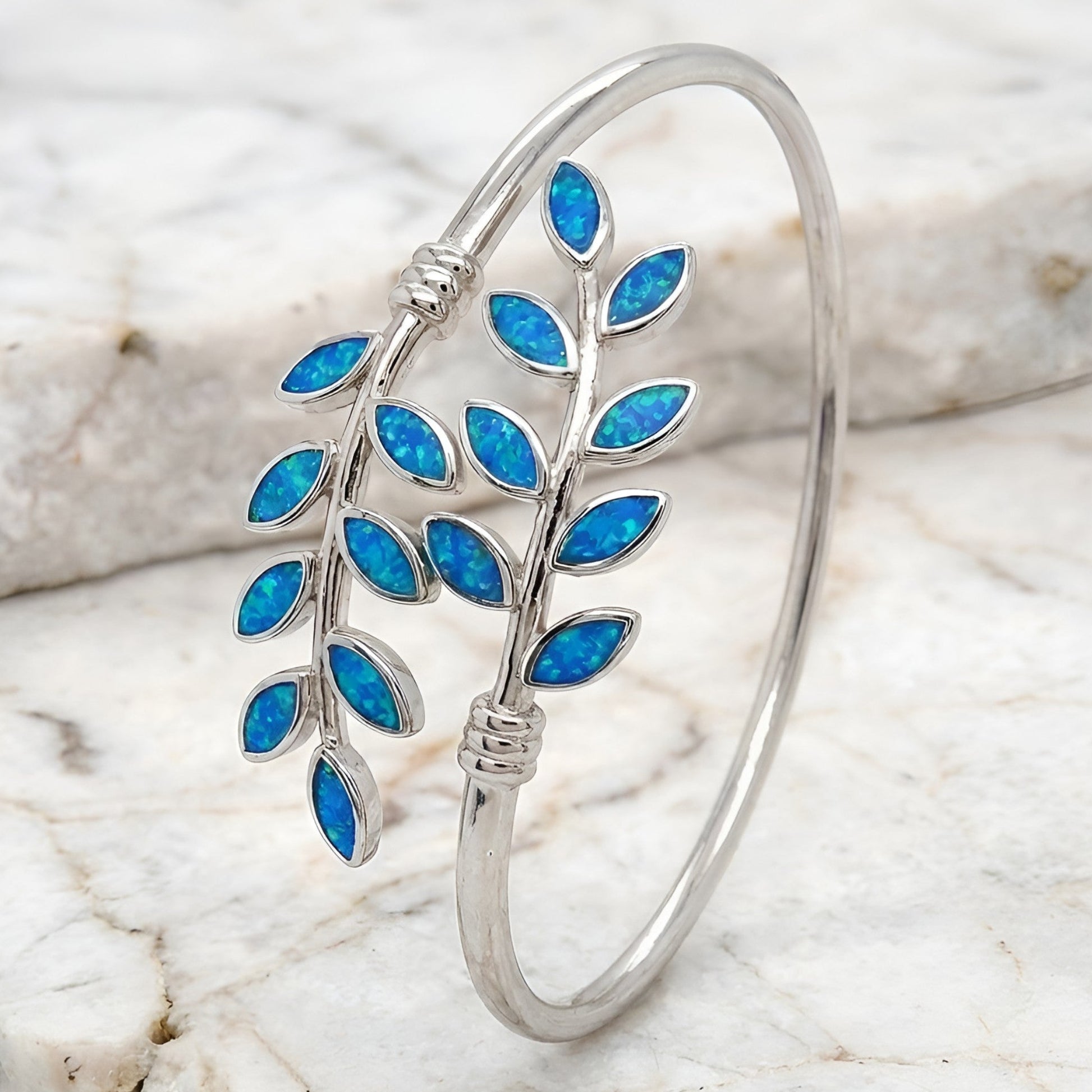 A silver open cuff bangle bracelet featuring a leafy branch design inlaid with vibrant blue opal-like leaves, displayed on a white marble surface.