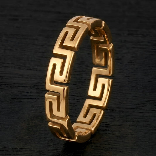 A gold Greek key pattern ring standing upright on a dark, textured background.