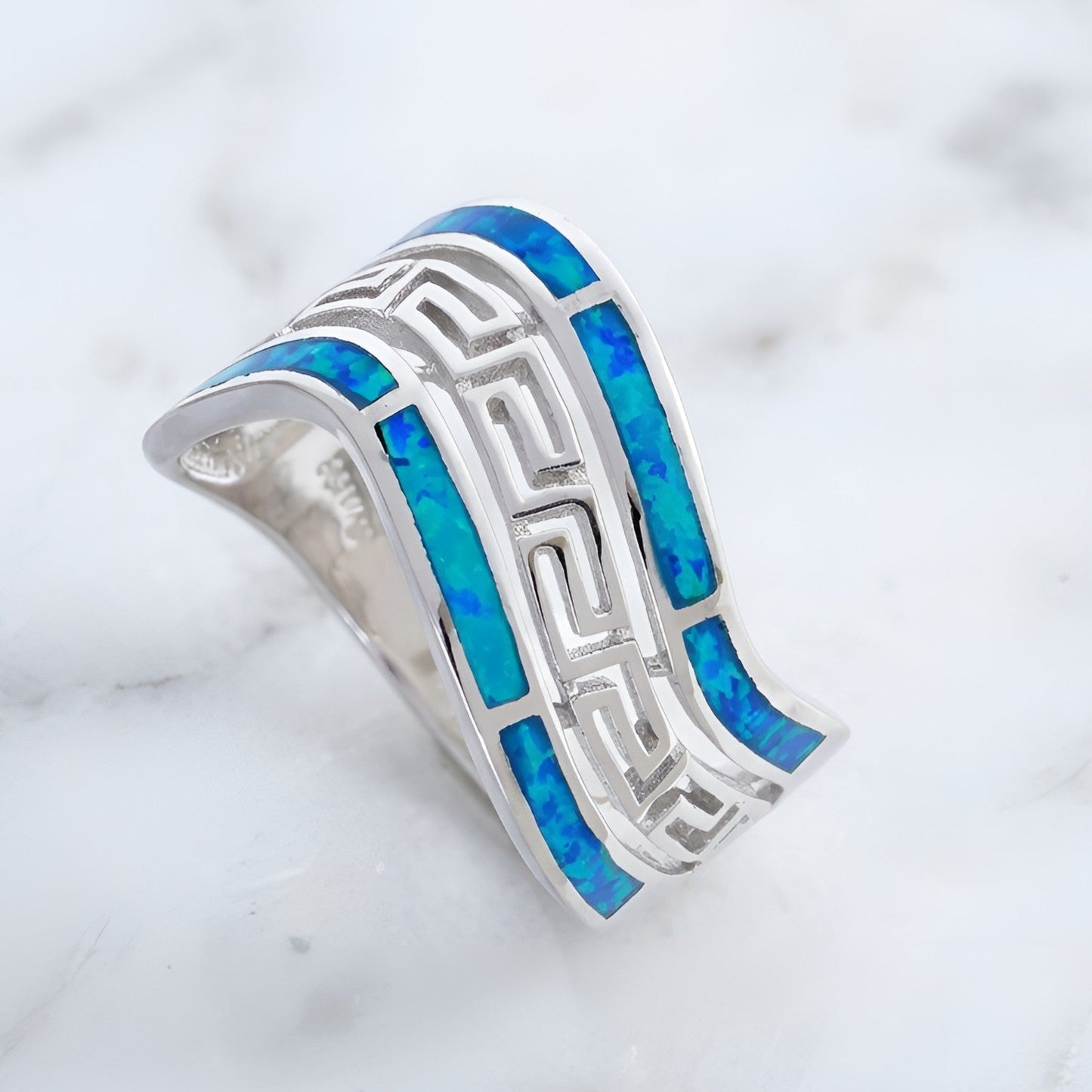 A silver Greek Key design band ring featuring vibrant blue opal inlay segments, photographed on a white marble surface.