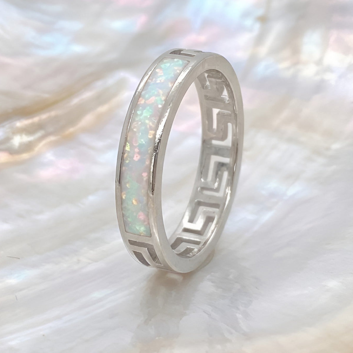A silver band ring with an inlay of iridescent white opal and a Greek key cutout pattern on the sides, displayed on a textured mother-of-pearl shell.
