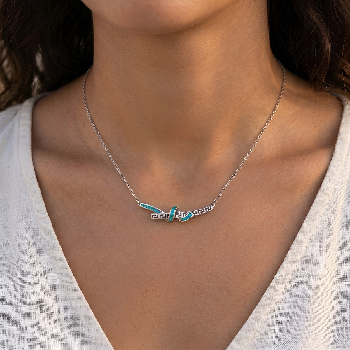 A close-up of a model wearing a silver chain necklace with a Greek key knot pendant featuring blue opal accents.