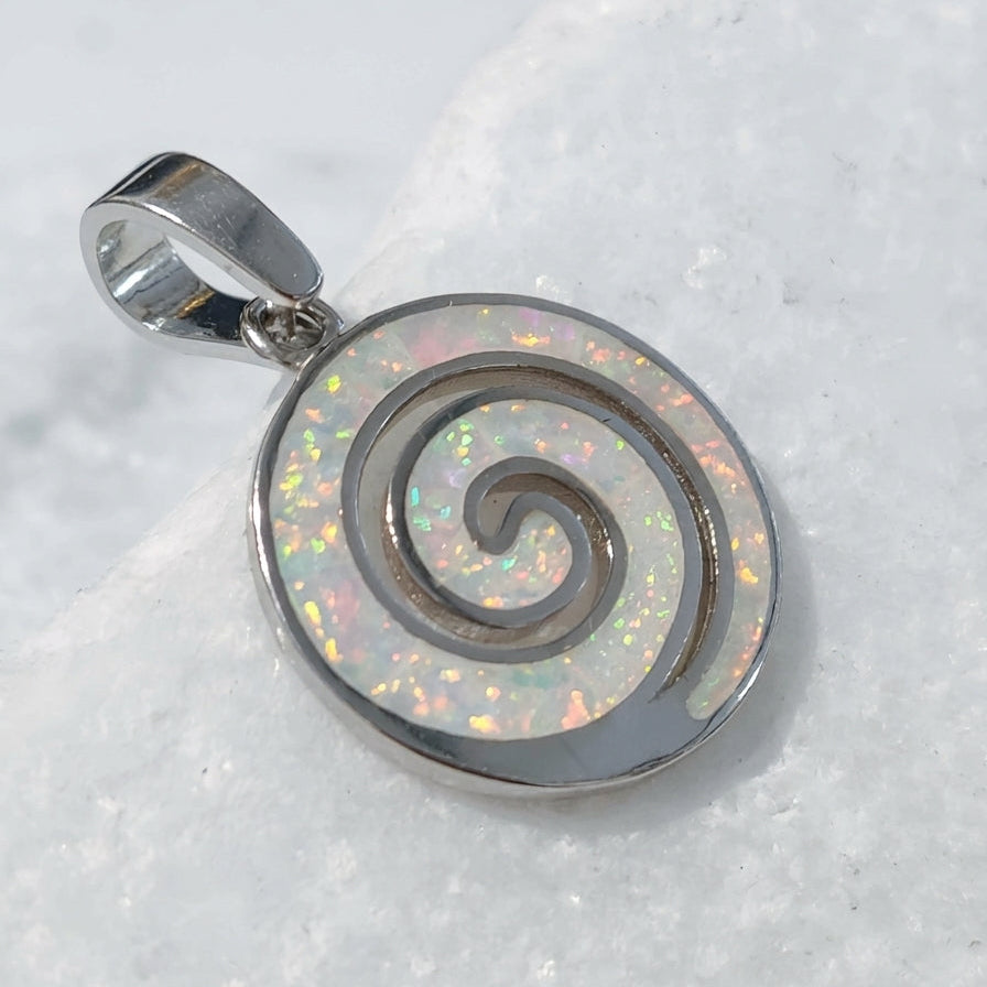 A silver spiral Greek pendant with an iridescent white inlay, resting on a textured white stone surface.