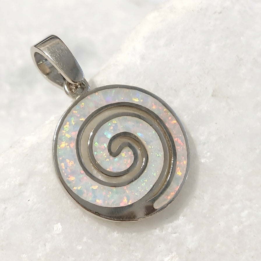 A silver spiral pendant with an iridescent white opal inlay, resting on a textured white stone surface.