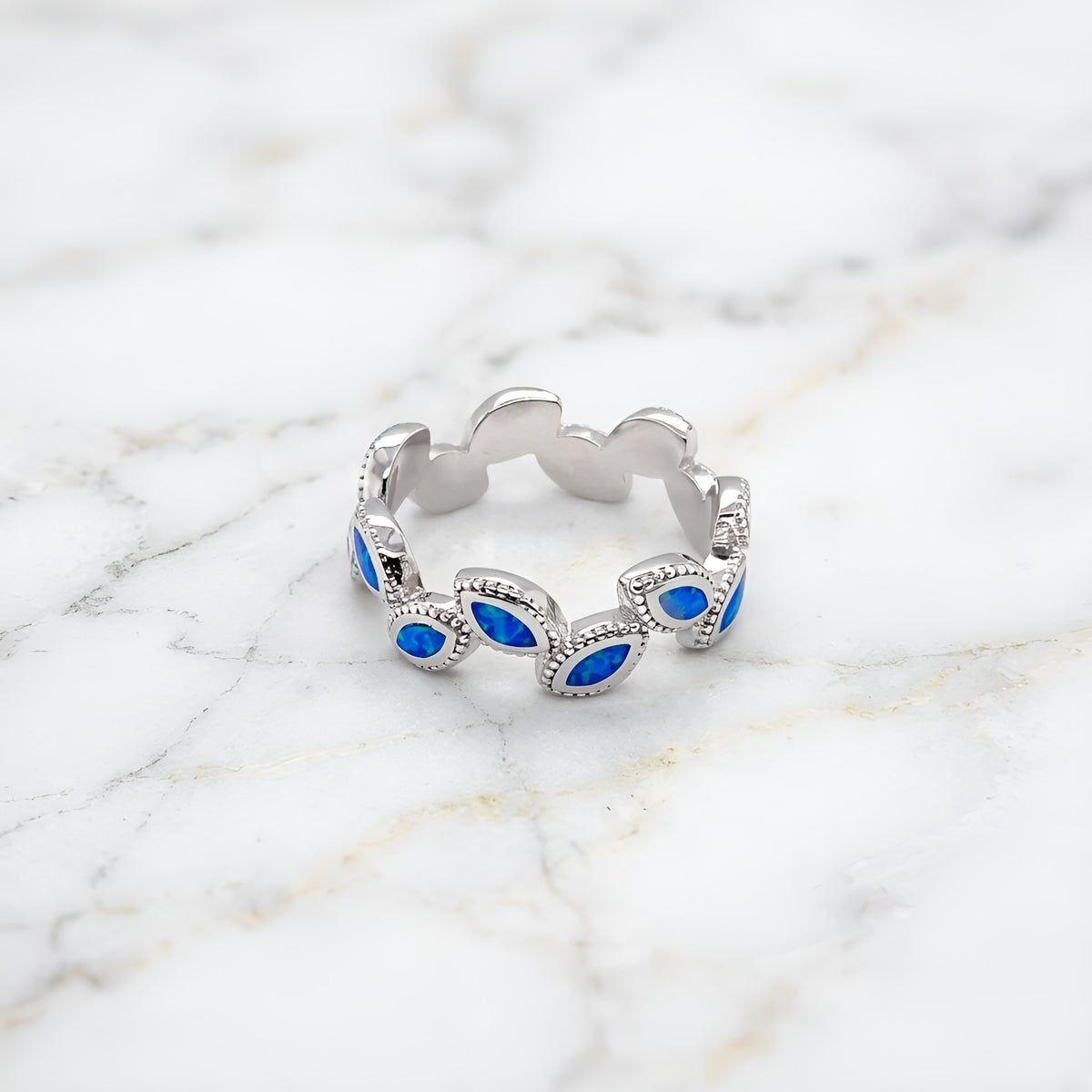 A silver band ring featuring a series of scalloped edges with marquise-cut blue stones, photographed on a white marble surface.