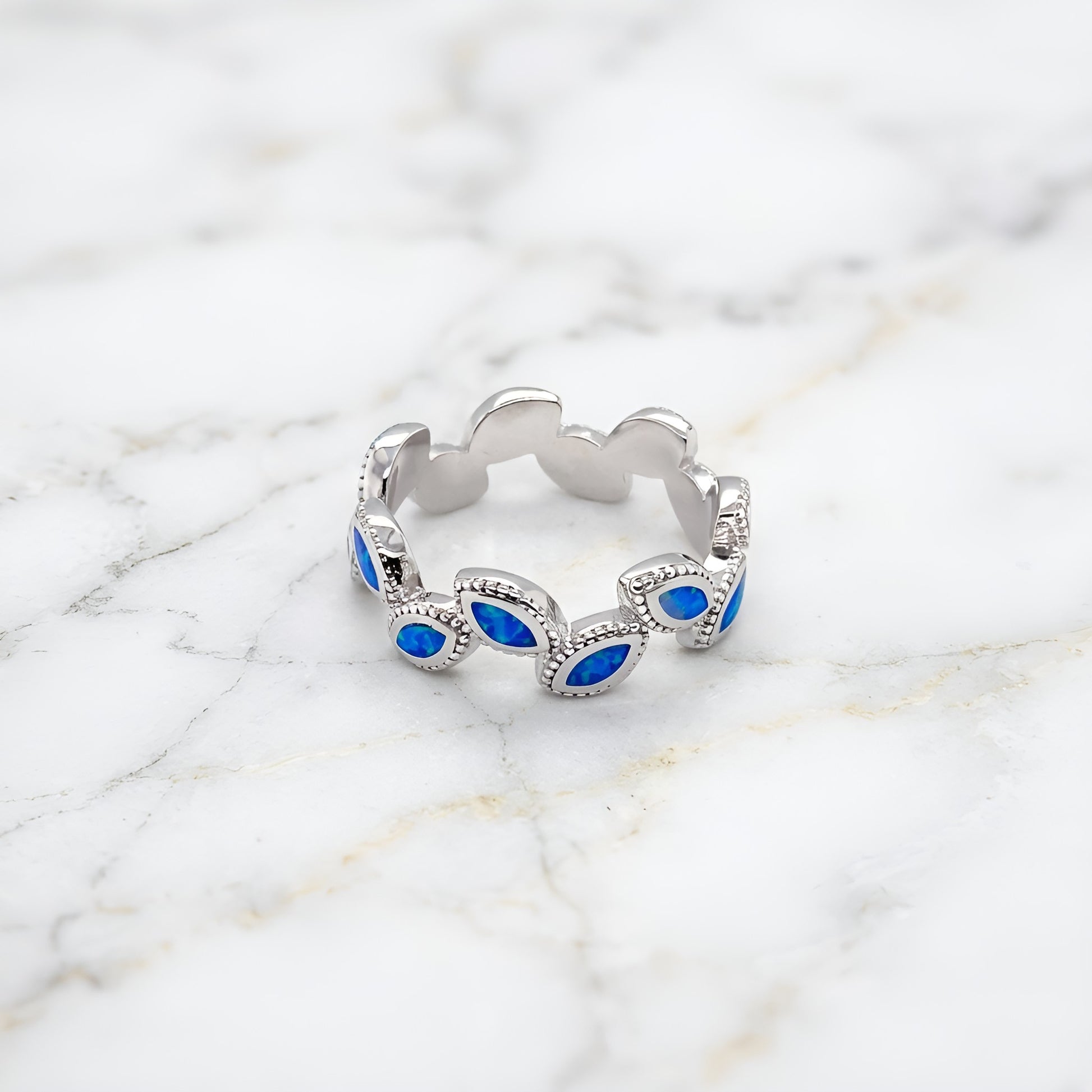 A silver band ring featuring a series of scalloped edges with marquise-cut blue stones, photographed on a white marble surface.