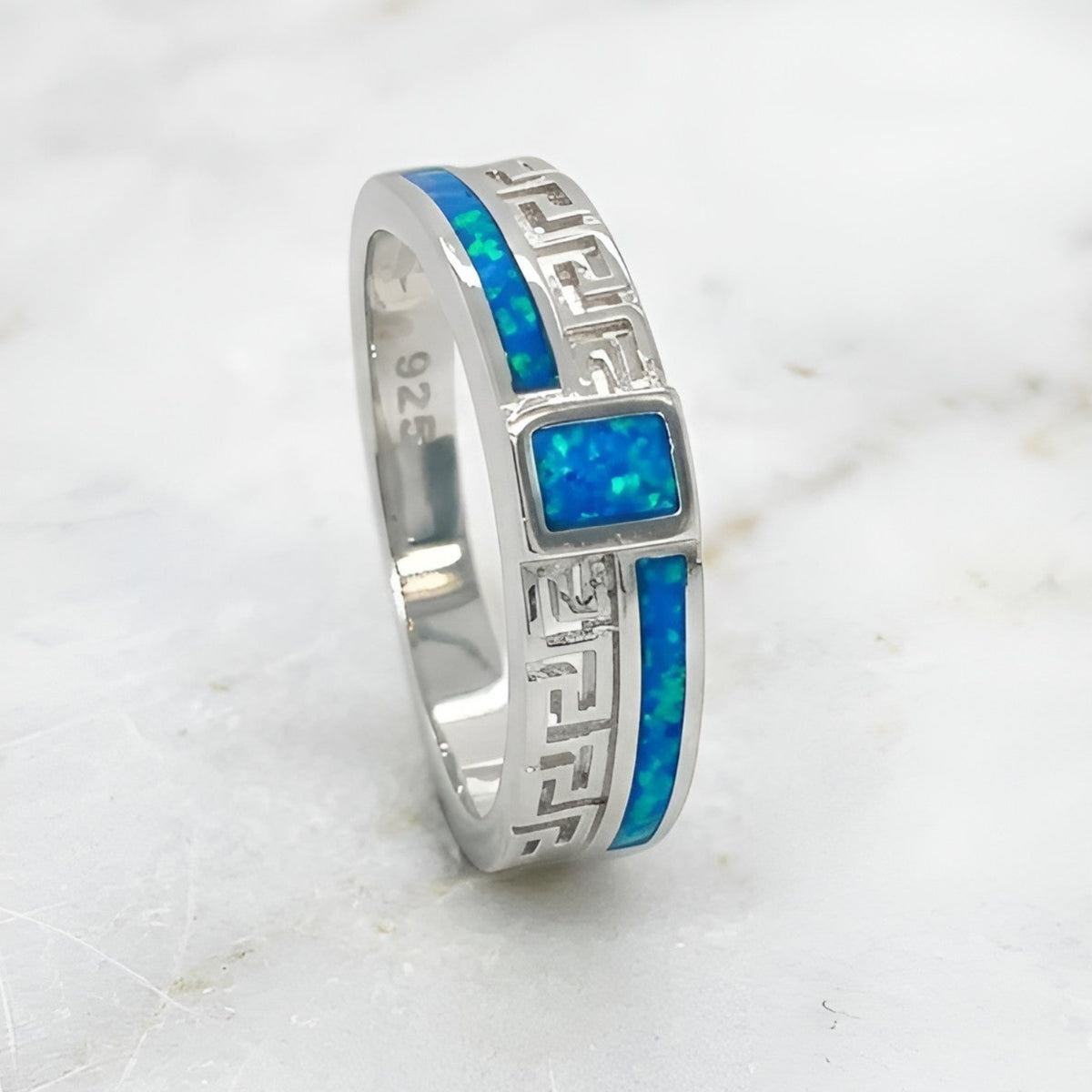 A silver band ring featuring a central rectangular blue opal stone and a side Greek key pattern inlaid with vibrant blue opal, displayed on a white marble surface.