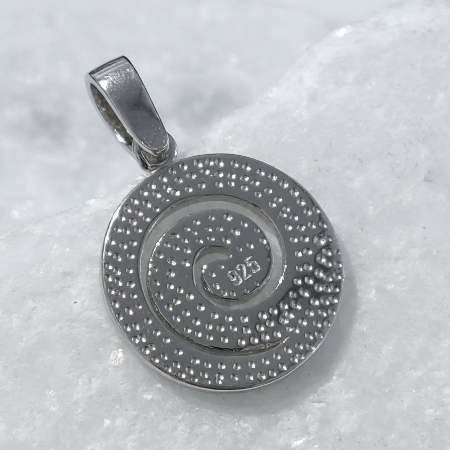 A silver spiral pendant with a textured beaded border and a polished center, resting on a white textured surface. The center features a '925' sterling silver hallmark.