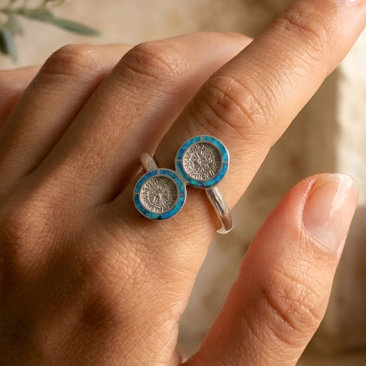 A close-up of a hand wearing a silver Phaistos Disc ring with two round blue and turquoise opal inlaid medallion-like tops, photographed in warm natural light against a softly blurred beige background.