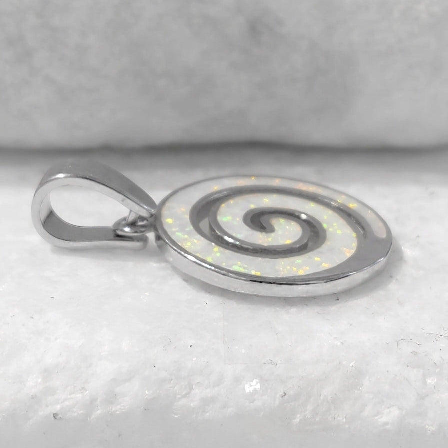 A silver Greek spiral pendant with an iridescent white inlay, resting on a textured white surface.