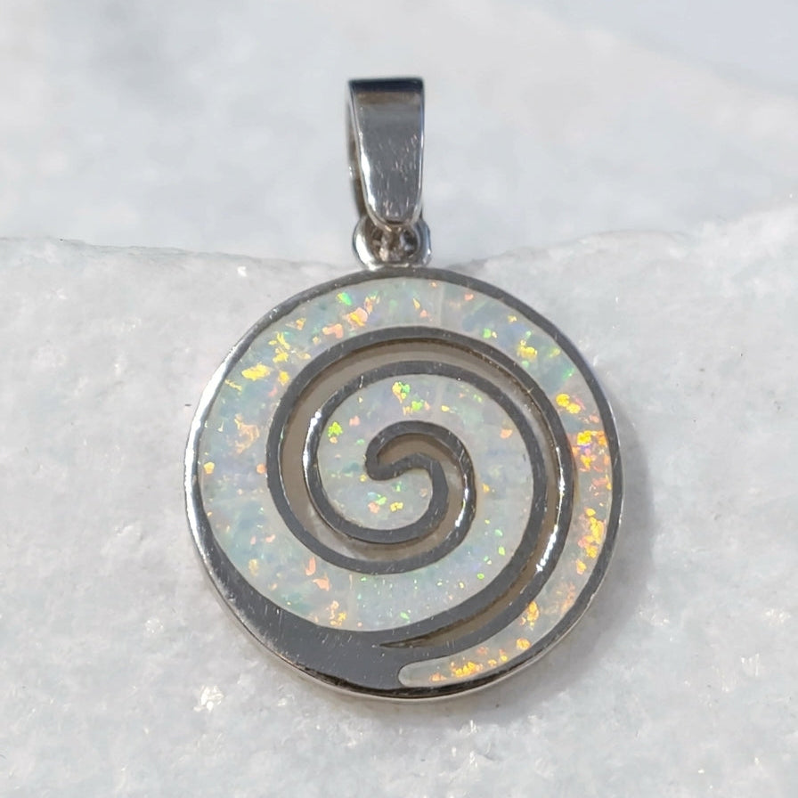 A Greek silver spiral pendant with an iridescent white opal inlay, resting on a textured white stone surface.