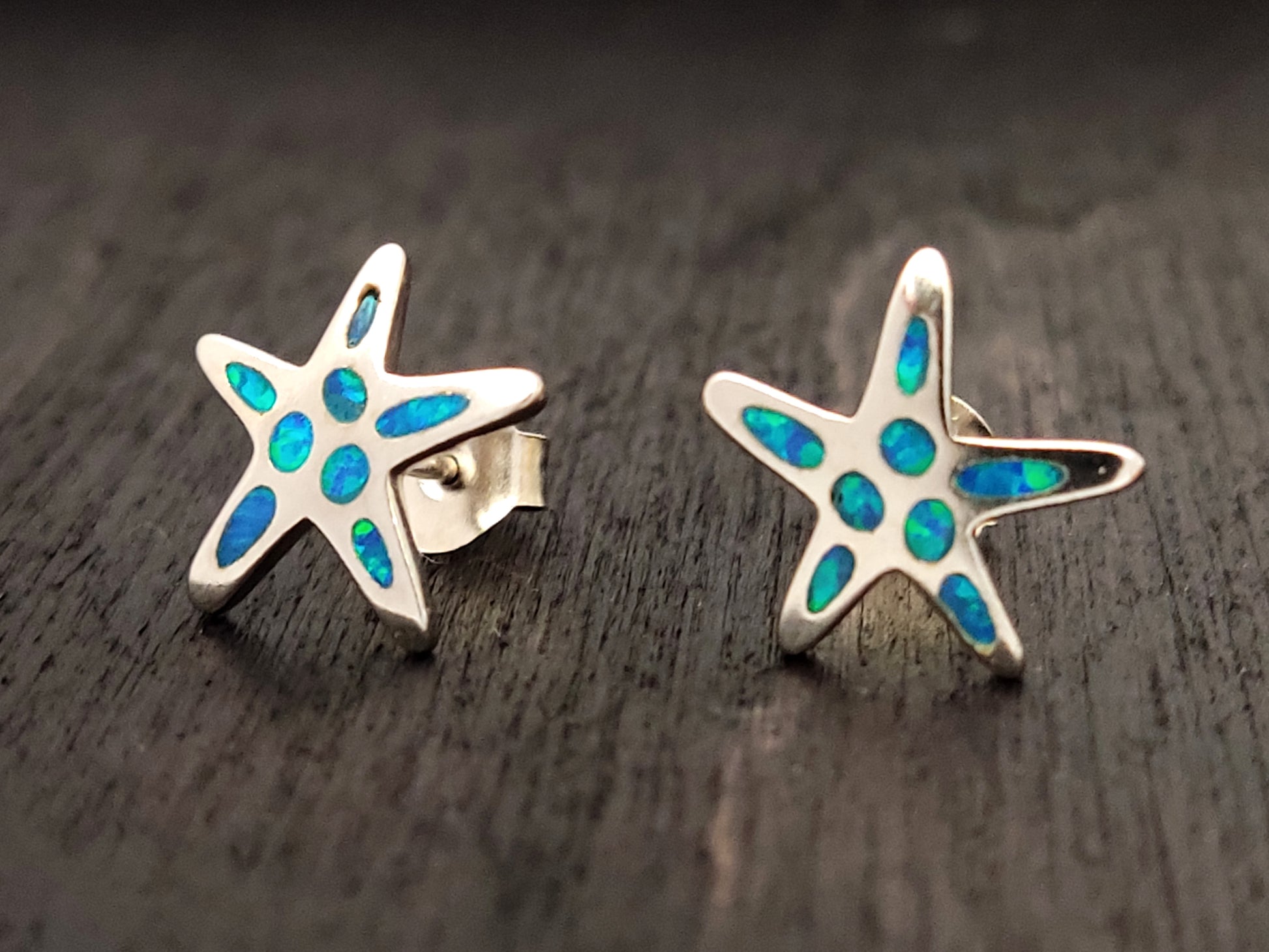 Starfish silver stud earrings with blue opal stones measuring 12mm on black background.