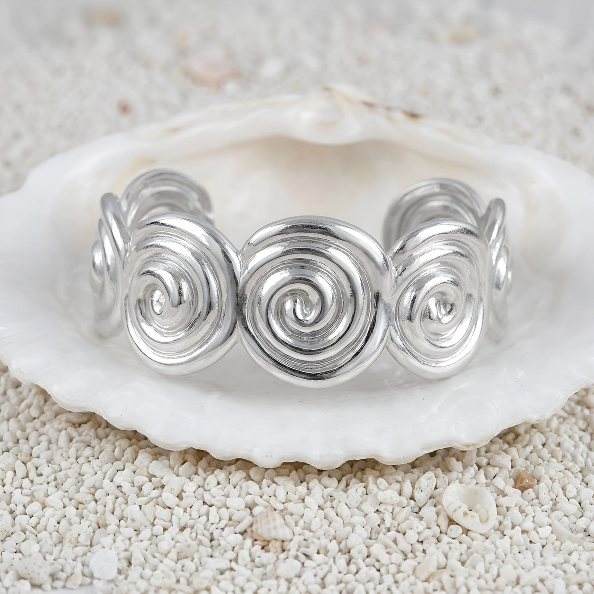 A silver spiral-patterned ring displayed on a white seashell over light sand.
