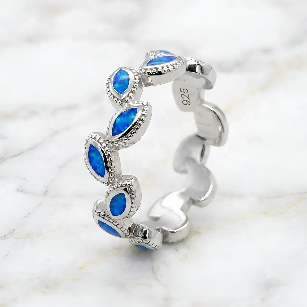 A sterling silver band ring featuring a series of marquise-cut blue opal inlays, photographed on a white marble surface. The inside of the band is stamped with '925' indicating sterling silver.
