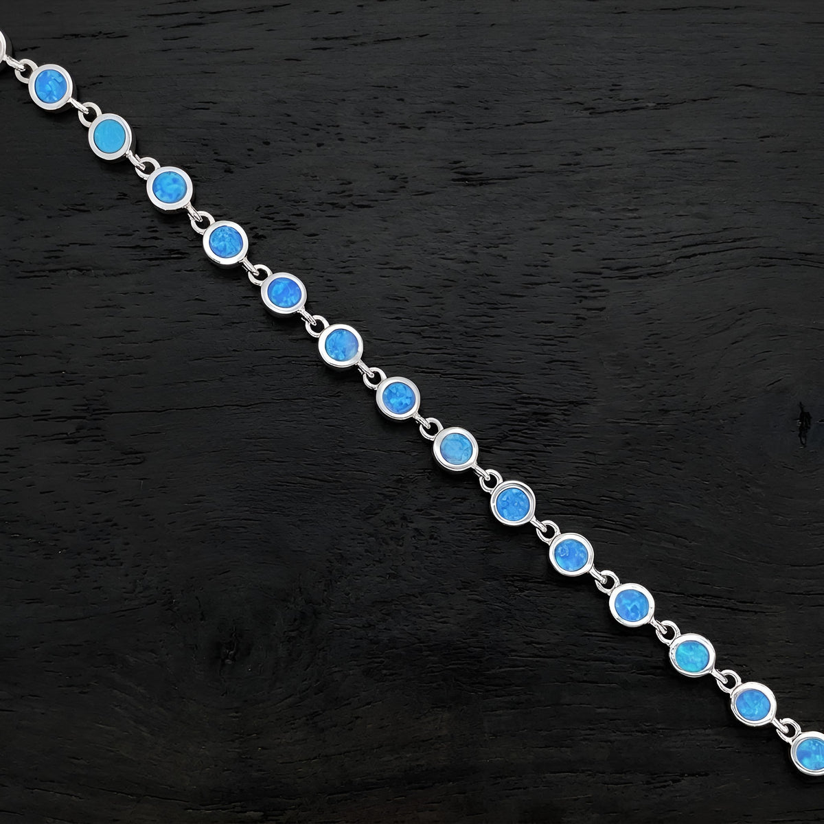 A silver chain bracelet featuring round, vibrant blue opal-like gemstones, displayed diagonally on a dark textured wooden surface.