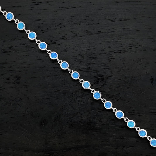 A silver chain bracelet featuring round, vibrant blue opal-like gemstones, displayed diagonally on a dark textured wooden surface.