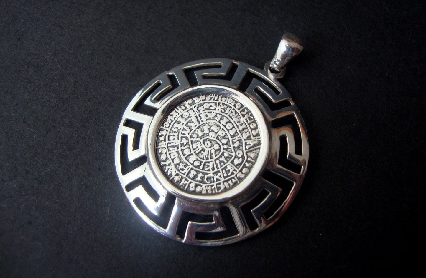 An intricately designed Sterling Silver 925 pendant featuring an Ancient Phaistos Disc & Greek Eternity Key Frame. The pendant has a diameter of 34 mm (1.32 inches) and a height of 40 mm (1.56 inches), including the hanger. Hallmarked 925. Handcrafted in Greece, showcasing rich cultural heritage and artisanal craftsmanship.